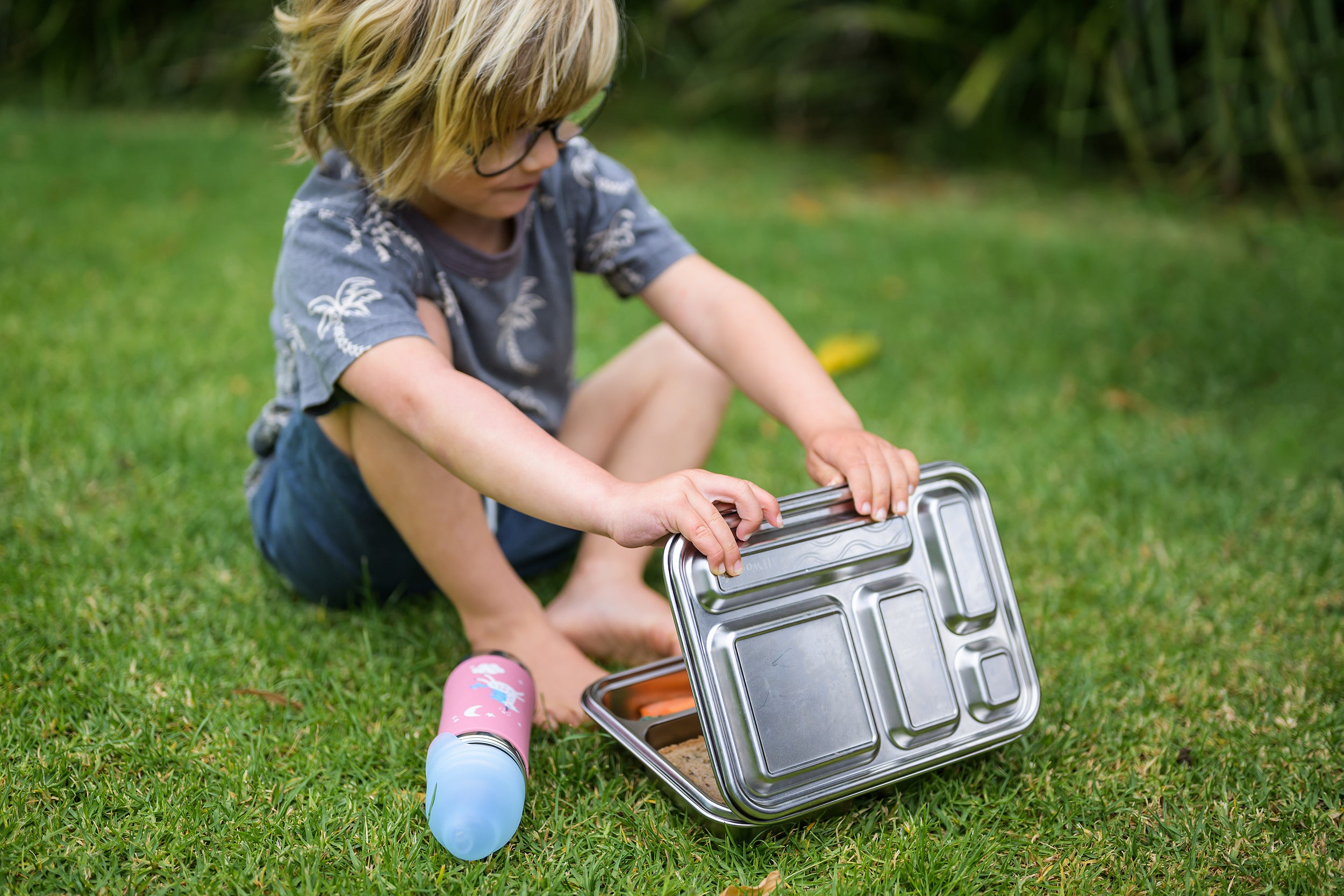 Boy sitting on grass opening a CaliWoods stainless steel bento lunchbox and a Pura toddler water bottle is on the ground next to it.