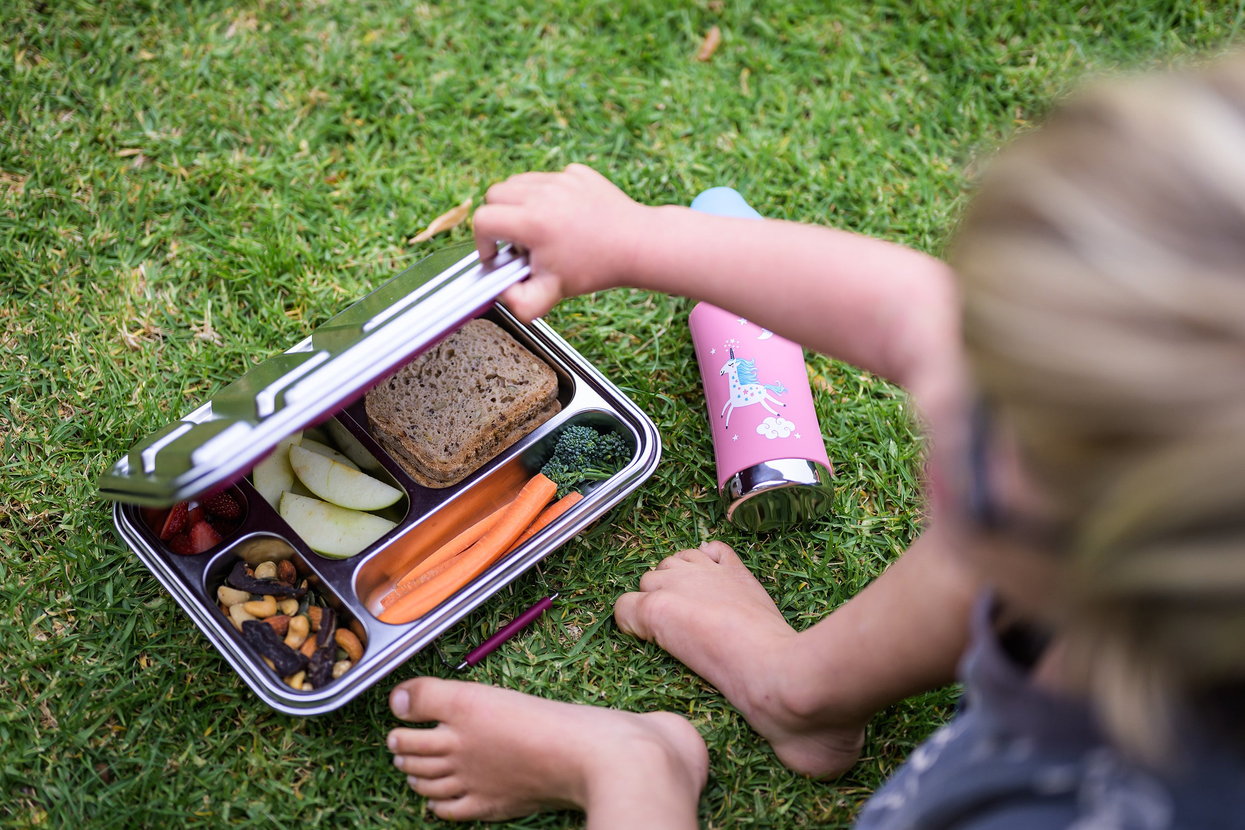 Over the shoulder shot of a little boy opening a stainless steel CaliWoods lunchbox with a Pura bottle next to it