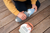 Stainless water bottle on a deck with a little boy opening the lid and a Silicone toddler snack box next to it