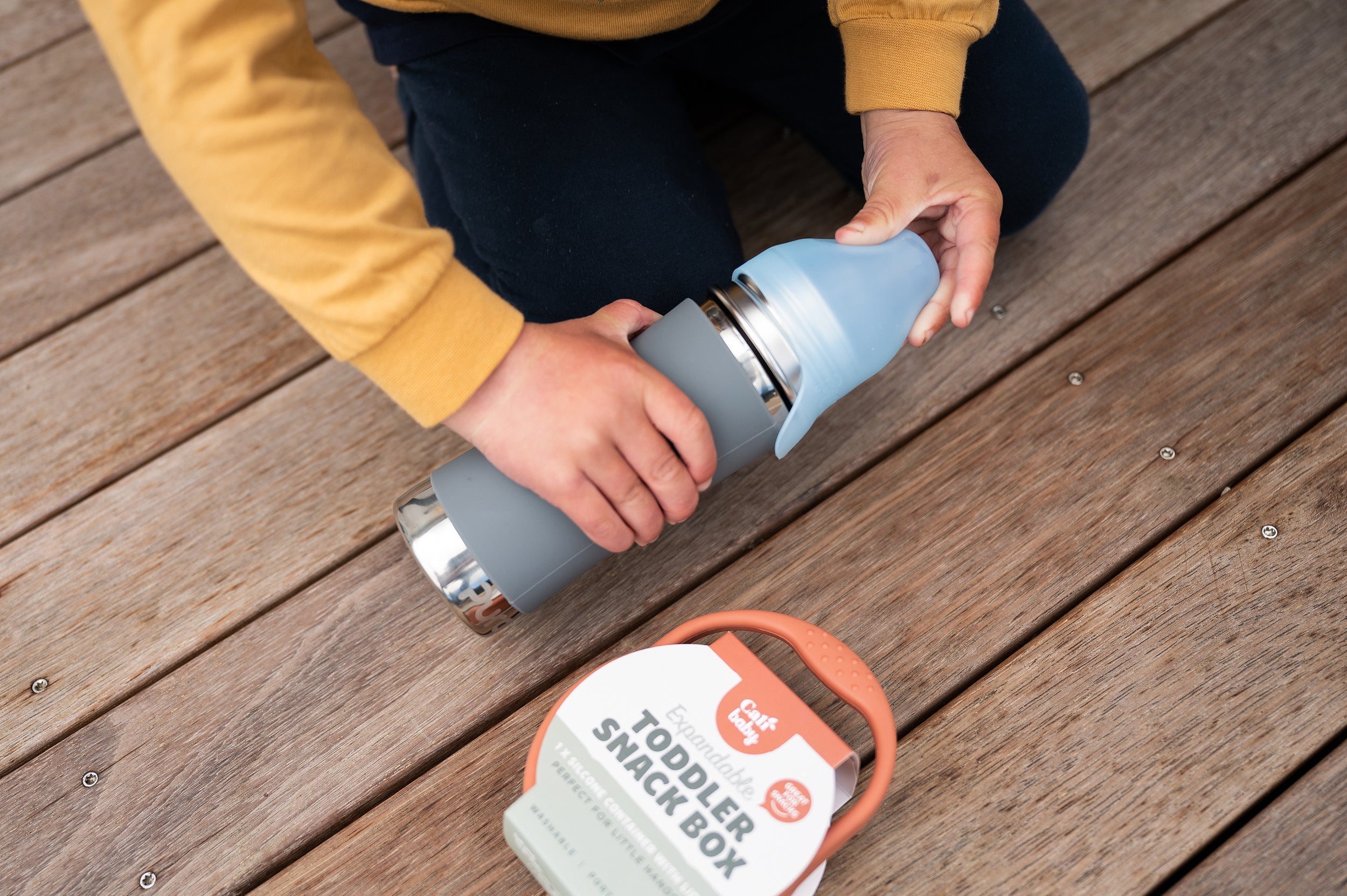 Stainless water bottle on a deck with a little boy opening the lid and a Silicone toddler snack box next to it