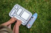 Little hands opening the CaliWoods Stainless bento lunchbox with toddler drink bottle on the grass