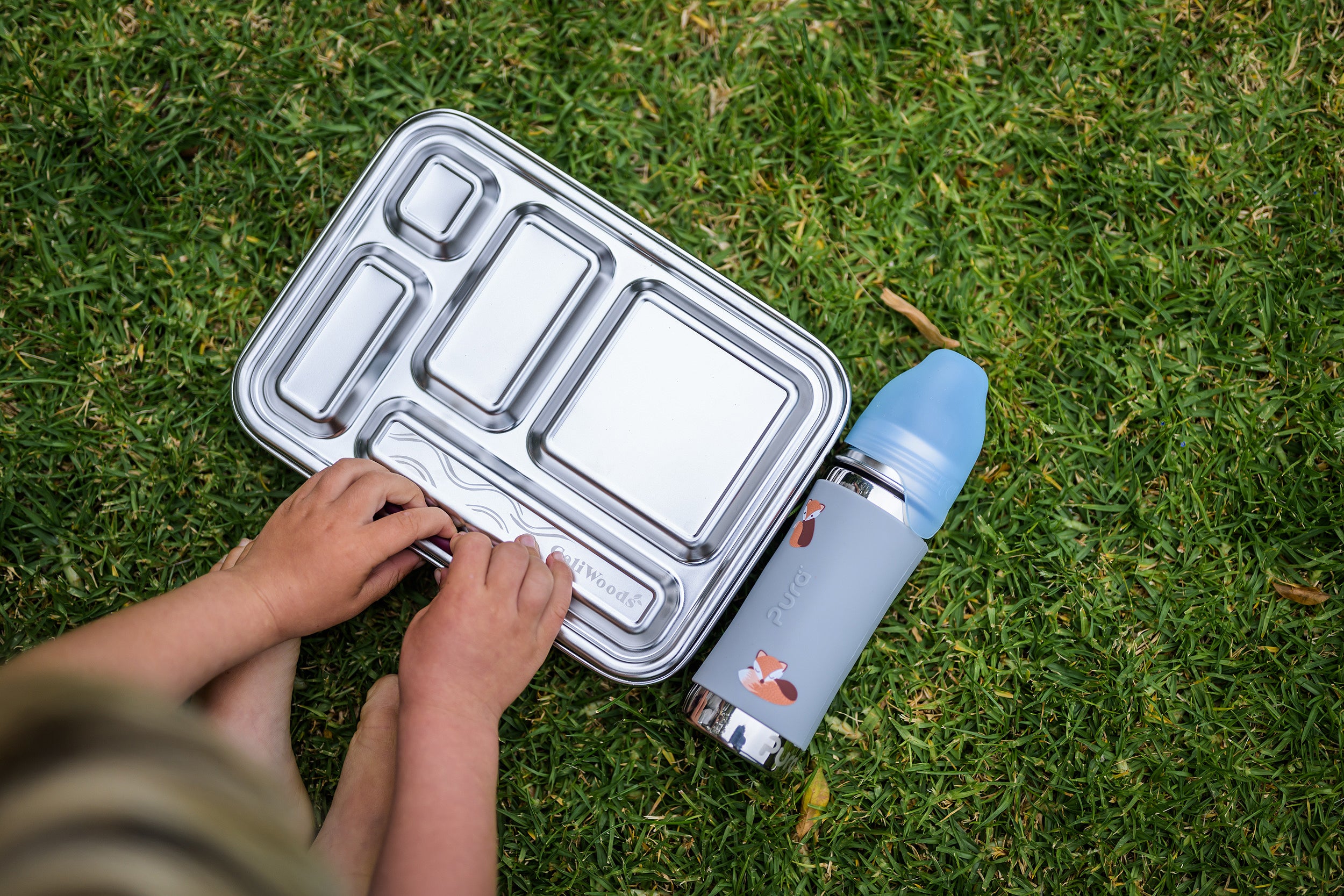 Little hands opening the CaliWoods Stainless bento lunchbox with toddler drink bottle on the grass