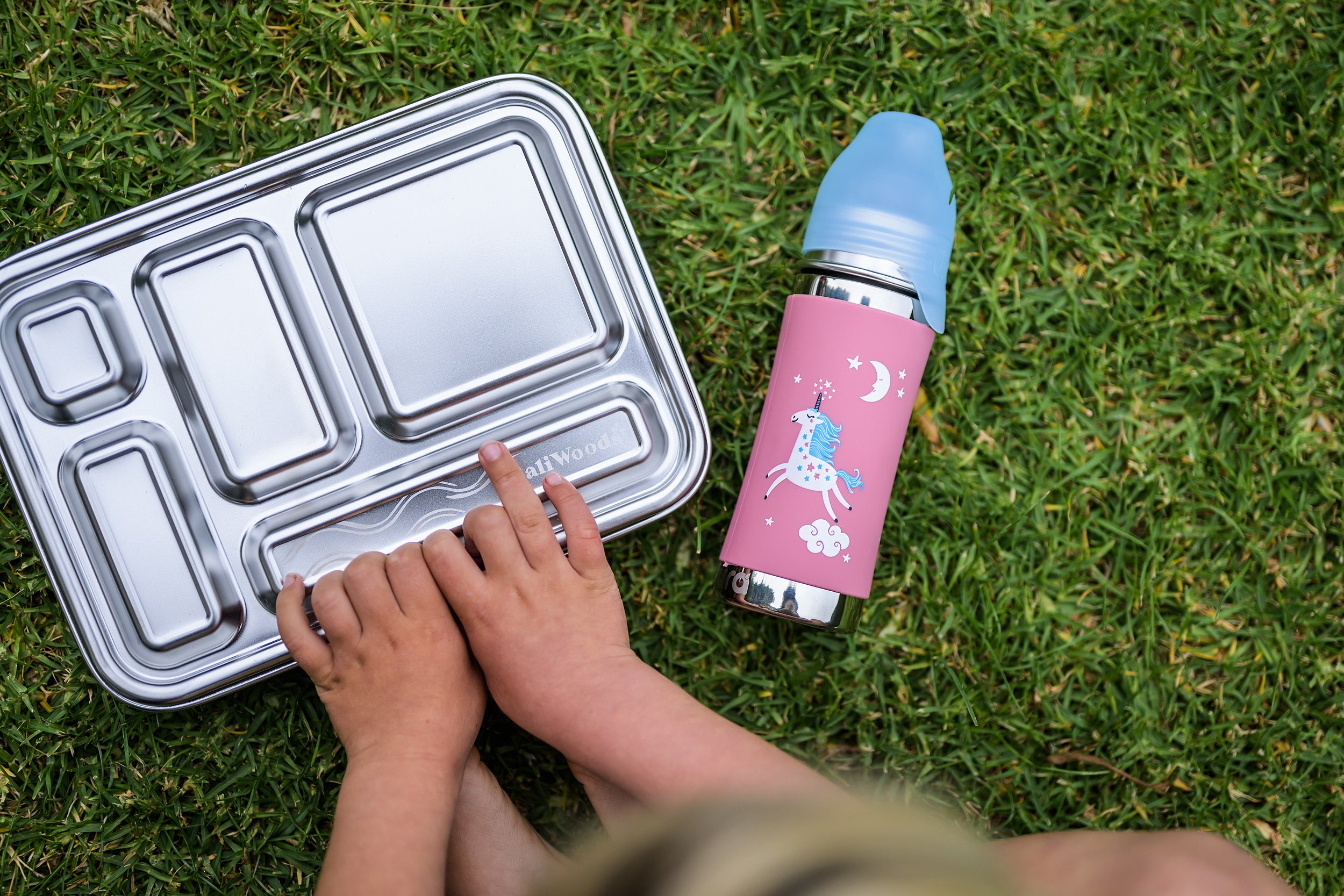 Pura Kiki toddler plastic free water bottle on the grass next to a CaliWoods stainless bento lunchbox. A little boy is using his hands to open the lunchbox 