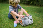 Boy sitting on grass opening a CaliWoods stainless steel bento lunchbox and a Pura toddler water bottle is on the ground next to it. 