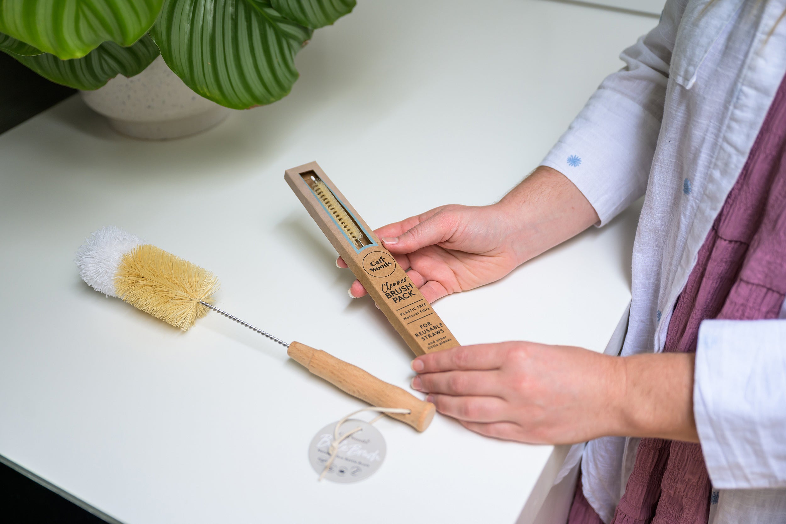 Hands holding the CaliWoods Cleaner Brush pack containing two natural straw pipe cleaners and Bottle Brush next to it on a kitchen bench. 