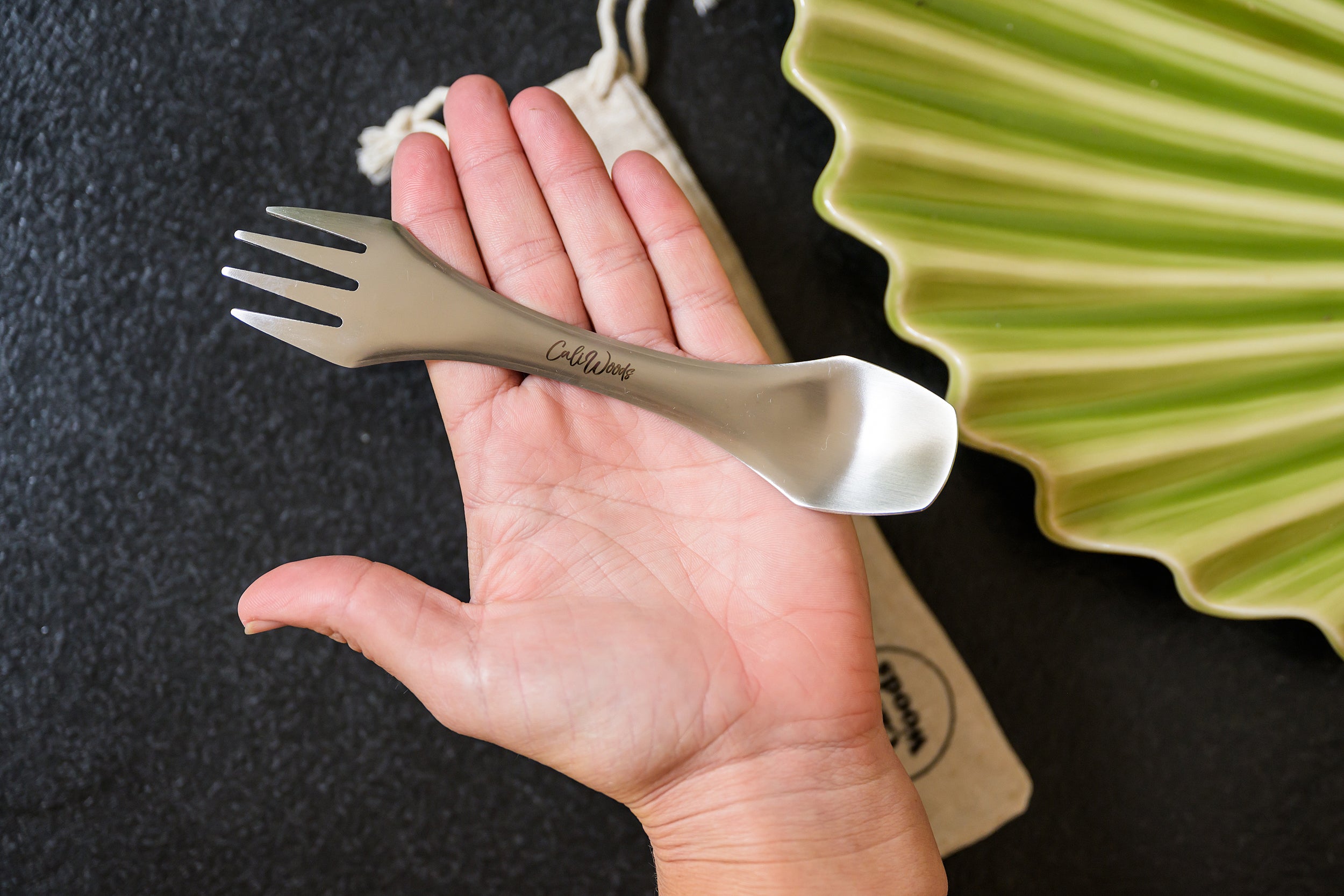 Stainless Spork on top of a flat hand with  linen carry bag in the back ground