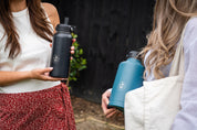 Close up of two reusable water bottles in hands, one blue bottle and one black bottle